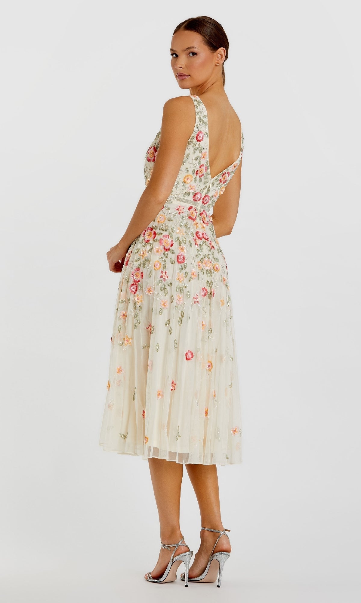 A woman models the Mac Duggal 5527 Floral-Embroidered Party Dress, a sleeveless cream tea-length dress, paired with silver high-heeled sandals, standing with her back turned and head looking over her shoulder at the camera.